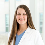 McKenna Steele, FNP-C, wearing a white coat over blue scrubs, smiling.